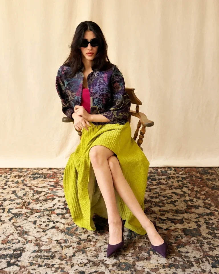Seated view of the Toranj Jacket and Skirt, emphasizing the form of the skirt, the textured patterned jacket, and the harmony of violet, fuchsia, and yellow-green tones
