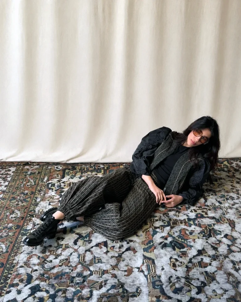 Reclining indoor view of the Fika Jacket and Trousers on a patterned carpet, emphasizing the shape of the trousers, the volume of the sleeves, and the dark tonal styling