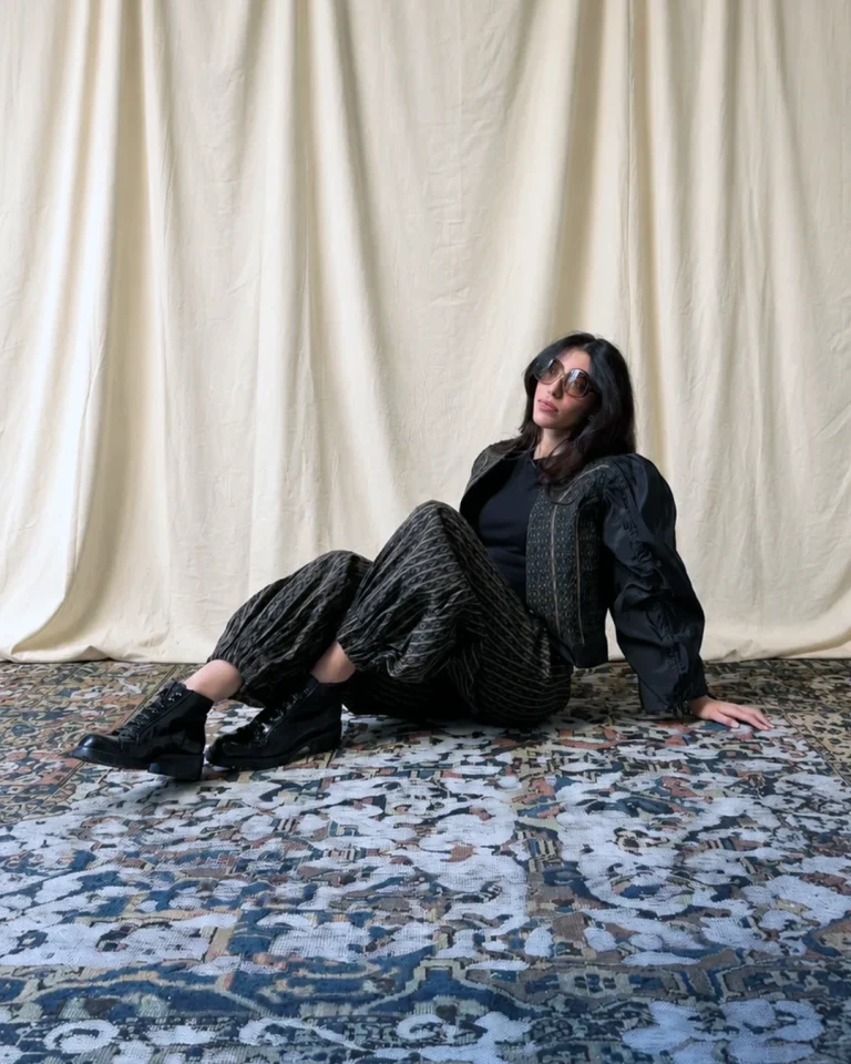 Seated indoor view of the Fika Jacket and Trousers, featuring a cropped textured jacket, patterned gathered-hem trousers, black boots, and sunglasses