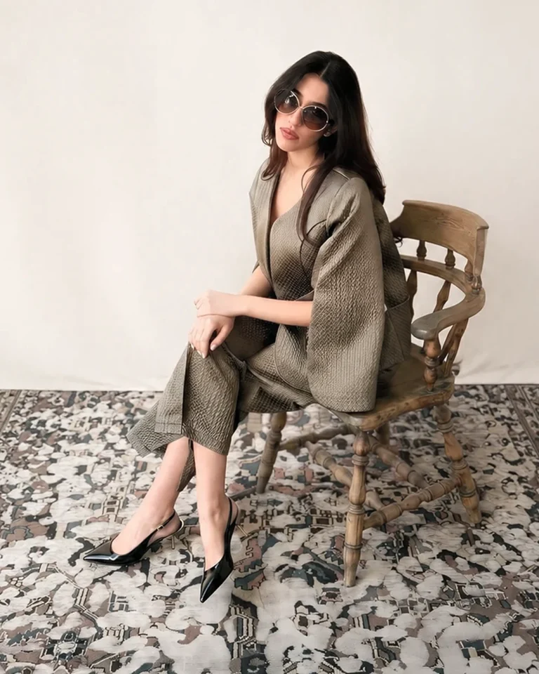 Seated indoor view of the Sedar Jacket and Trousers on a wooden chair, featuring a short relaxed jacket, straight trousers, and black heels