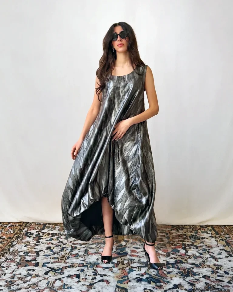 Full-length view of the Silver Silk Dress with a free voluminous silhouette, luminous waved surface, and black heeled sandals indoors