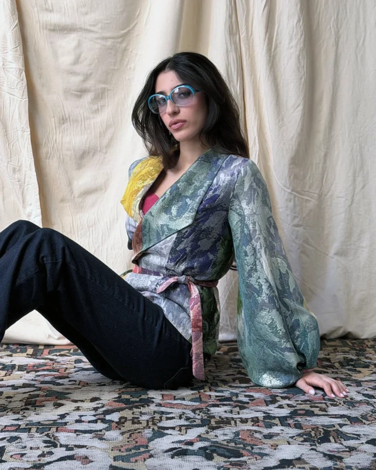 Seated view of the Zarvan Handwoven Jacket, featuring Atlasi Daraei and Zari weaving, voluminous sleeves, a fabric belt, and dark trousers
