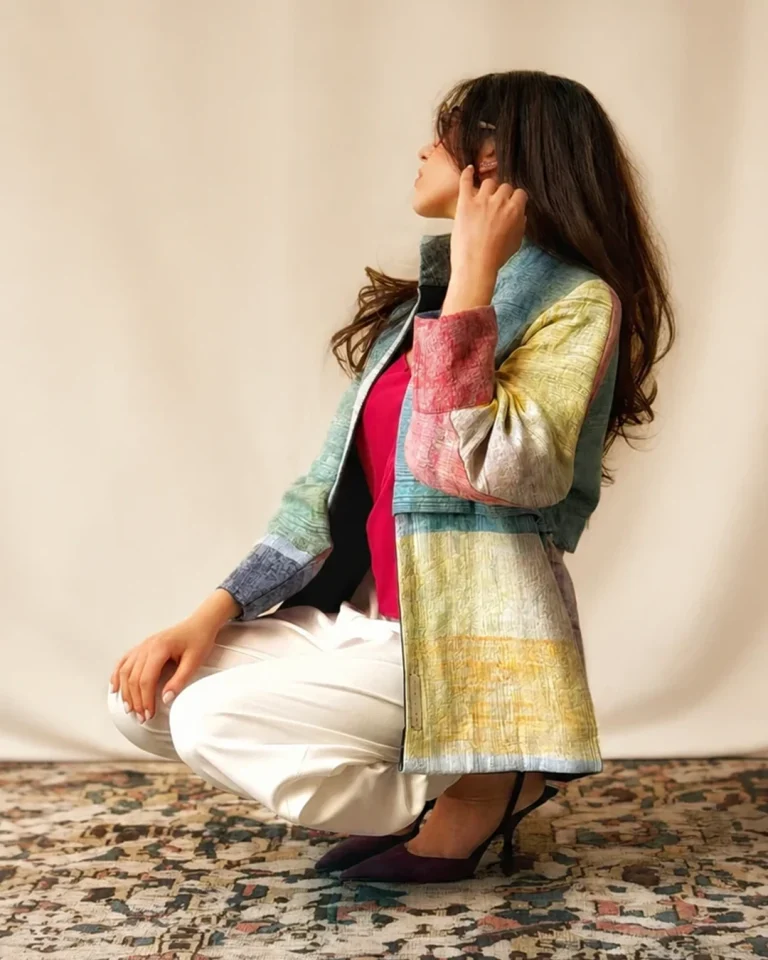Seated model wearing the Iron Jacket in multicolored handwoven Daraei cotton with a fuchsia top, light trousers, and dark heels
