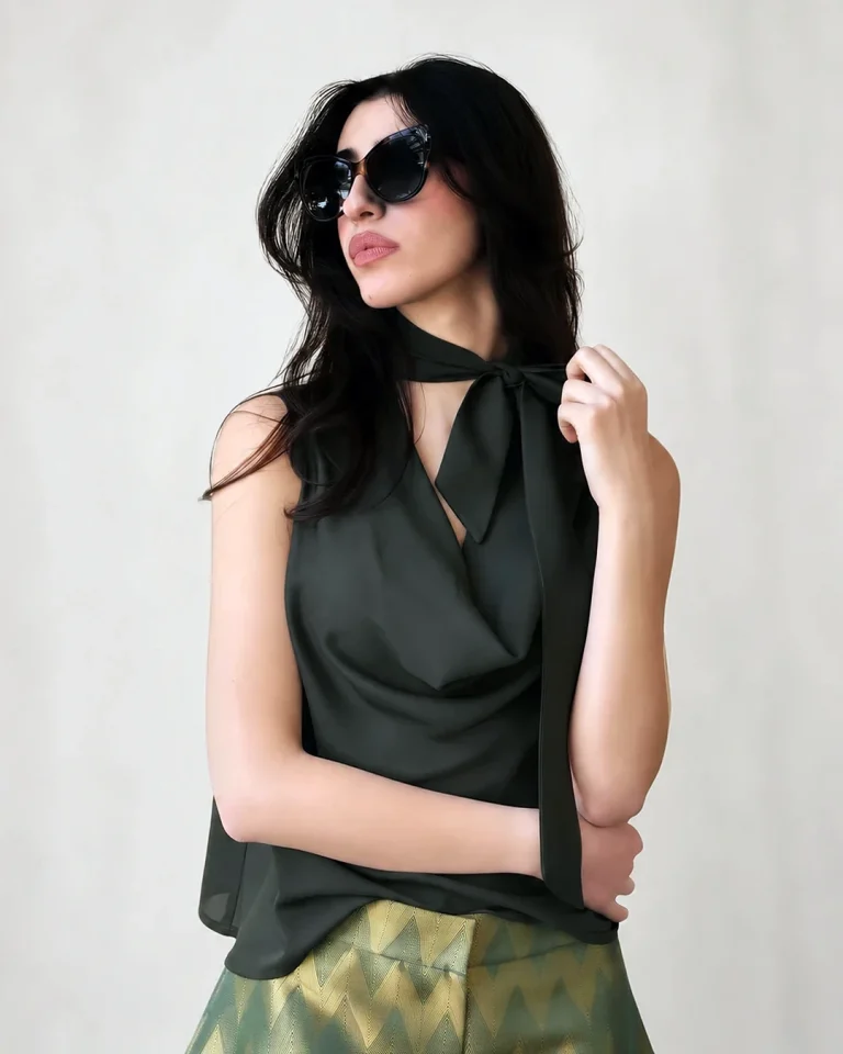 Portrait of a model wearing the dark green Zarrin blouse with a long tie-neck detail, sunglasses, and green-gold patterned trousers