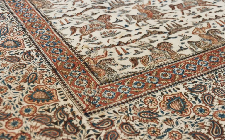 Close-up of a hand block-printed Persian ghalamkar textile from Isfahan, showing intricate hunting and polo-playing scenes with horsemen printed in rust-red and blue on cream cotton, framed by floral borders and paisley (boteh) motifs, a traditional craft made with hand-carved wooden stamps and natural dyes.