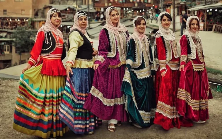 Six Iranian women posing in vibrant traditional Gilaki clothing in the historic stepped village of Masuleh, Gilan, wearing brightly colored ribbon-trimmed pleated skirts (Shaliteh), silver-braided velvet vests (Jalizgheh), and netted lace headscarves (Lachak).