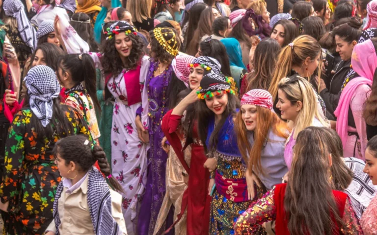 A large crowd of Kurdish women and girls joyfully dancing in traditional attire during a celebration in western Iran, wearing richly colored and sequined long dresses, embroidered vests, fringed floral headdresses (Kurdish Klash/Jamaneh wraps), and coin headbands.