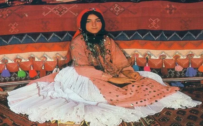 A young Qashqai woman seated on a handwoven Persian rug, dressed in traditional attire consisting of a voluminous layered white ruffled skirt, a pink sequined tunic with coin necklace embellishments, and a red headscarf, backed by vibrant tribal kelims and hanging tassels.