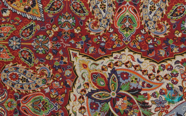 Close-up of Persian termeh fabric in rich crimson red, intricately woven with teardrop-shaped boteh (paisley) motifs, floral sprays, and a central medallion (Toranj) design in gold, cream, blue, and green silk threads — a traditional textile craft of Yazd, Iran.