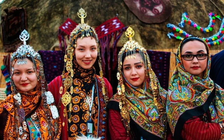 Four Turkmen women in northeastern Iran wearing traditional red and striped silk tunics, large floral and paisley headscarves (Yalyk), and elaborate tall silver and gold-plated forehead crowns (Gupba) adorned with dangling pendants, standing in front of a traditional nomadic yurt.