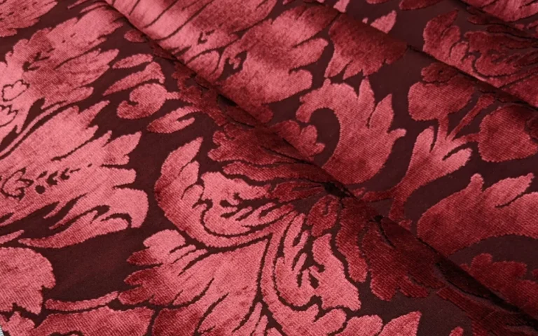 Close-up of luxurious Persian cut velvet fabric in rich crimson and maroon tones, featuring embossed large-scale floral and acanthus leaf patterns with a lustrous sheen, representing traditional Iranian luxury textiles historically rooted in Safavid-era formal wear.